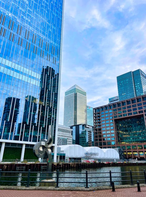 A cityscape featuring several modern high-rise buildings with reflective glass facades, including a prominent tall tower on the left with vertical linear patterns. In the foreground, a body of water is bordered by a black metal railing, with a large metal sculpture resembling intertwined loops positioned close to the water’s edge. Behind the sculpture, there are white, tent-like structures on the pavement, alongside a variety of commercial office buildings with different architectural styles, materials, and finishes. The sky above is clear with some light cloud cover, providing natural daylight that highlights the glass surfaces of the buildings. The scene suggests an urban environment with active development, and the presence of structures and surroundings associated with city employment and business districts, reflecting typical features of a financial or commercial area where private waste handling and independent rubbish removal services could be relevant.