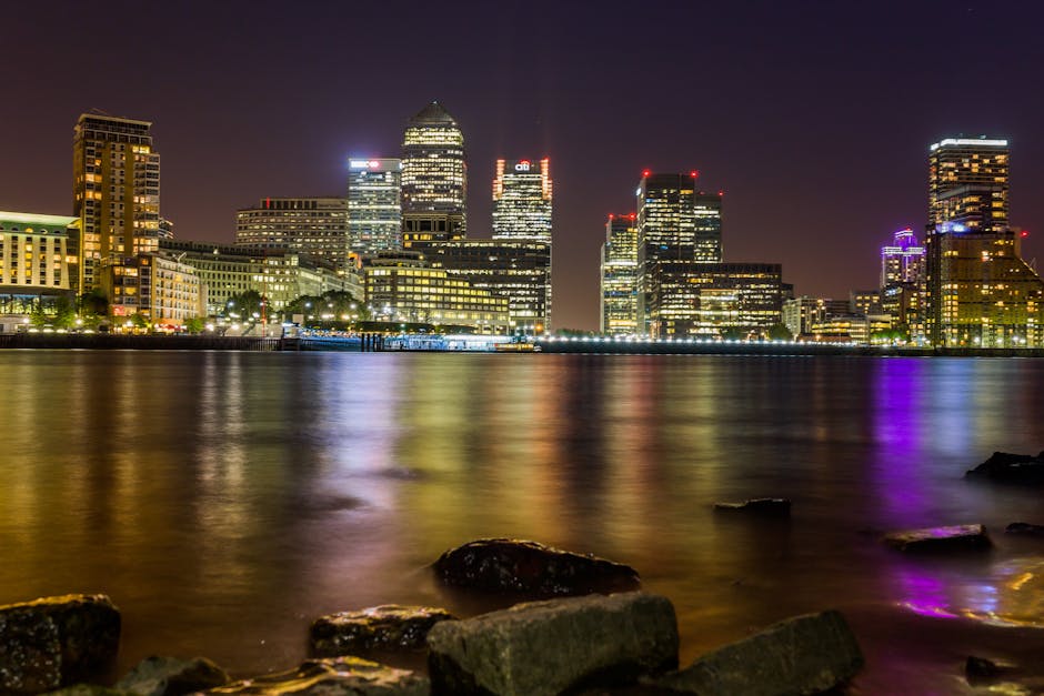A nighttime cityscape of a modern financial district with illuminated high-rise buildings reflected in the calm water of a river or harbour. In the foreground, dark rocks and stones are visible along the shoreline. The buildings vary in height and architecture, with some topped by distinctive lighting or signage, and their glass facades reflect vibrant multicoloured lights, including purple, blue, and white. The scene is captured under clear skies, with the city lights creating a bright, dynamic atmosphere. This urban environment showcases the contrast between the natural waterfront and the dense, lit-up skyscrapers, subtly aligning with ideas of private waste handling and alternative collection methods for urban debris or rubbish clearance, reminiscent of independent waste management in busy city areas. The overall composition emphasizes the clean, reflective water surface and the detailed textures of the rocks in the foreground, providing a professional and factual visual suitable for a rubbish removal blog or service-related content relating to city waste management.