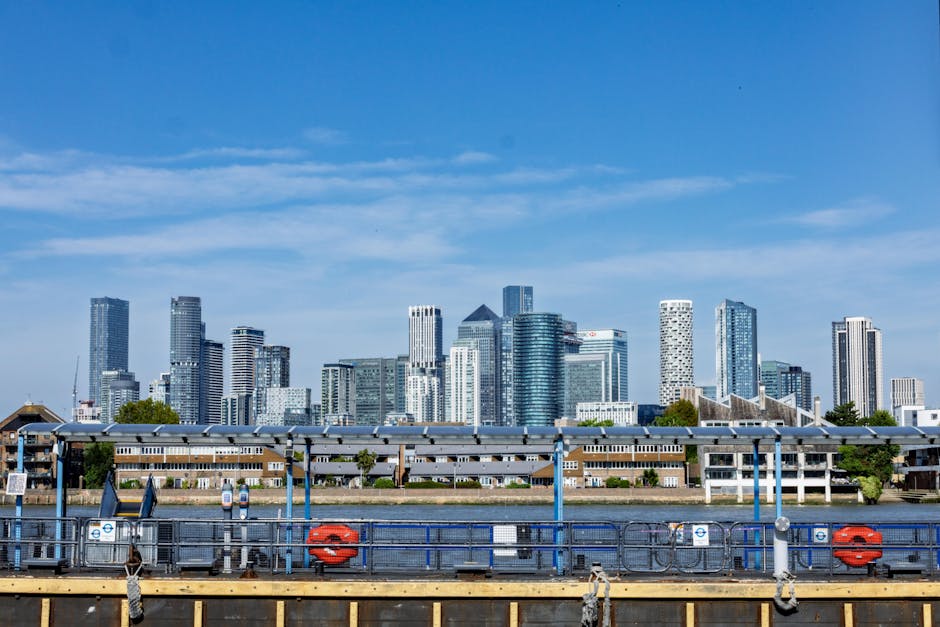 A cityscape featuring several modern high-rise buildings with reflective glass facades, including a prominent tall tower on the left with vertical linear patterns. In the foreground, a body of water is bordered by a black metal railing, with a large metal sculpture resembling intertwined loops positioned close to the water’s edge. Behind the sculpture, there are white, tent-like structures on the pavement, alongside a variety of commercial office buildings with different architectural styles, materials, and finishes. The sky above is clear with some light cloud cover, providing natural daylight that highlights the glass surfaces of the buildings. The scene suggests an urban environment with active development, and the presence of structures and surroundings associated with city employment and business districts, reflecting typical features of a financial or commercial area where private waste handling and independent rubbish removal services could be relevant.