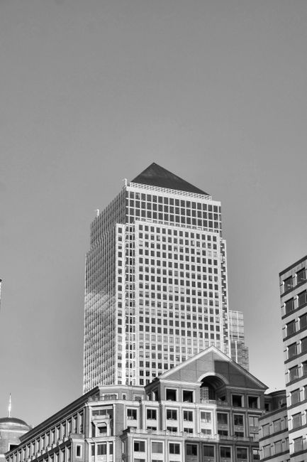 A black and white photograph of a modern high-rise office building with a rectangular shape and a pointed roof, featuring a grid of large windows covering its facade. In the foreground, lower-rise historic-style buildings with brick or stone exteriors, detailed window frames, and pitched roofs are visible, contrasting with the taller structure behind them. The scene is set in an urban environment with clear, bright lighting that highlights the textures of the building materials. The area appears to be in a city centre, possibly in Canary Wharf, with the high-rise contributing to the skyline. The photograph emphasizes the architecture and the arrangement of buildings, subtly reflecting the context of urban property development and alternative disposal methods where businesses may arrange private waste collection for such locations. The overall scene captures the mix of historical and modern architectural styles typical in city centres with prominent commercial buildings.