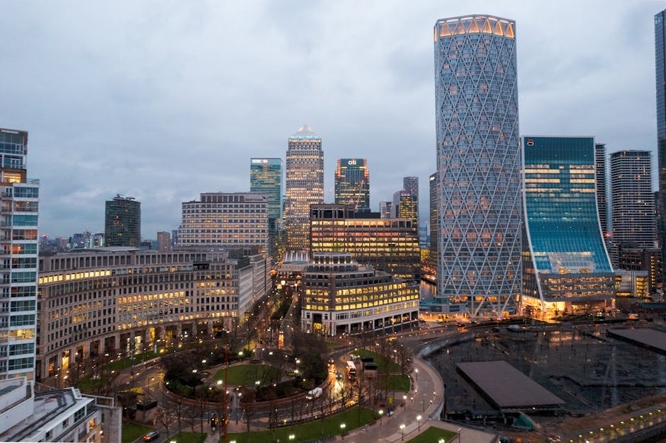 A cityscape view of a modern urban area during dusk, featuring a variety of high-rise office buildings with distinct architectural designs and illuminated windows. The tallest building has a crisscross diamond-patterned facade with a rounded top, situated prominently on the right side of the image. Surrounding buildings display glass exteriors reflecting the evening sky and city lights, creating a vibrant skyline. In the foreground, there is a park with a circular pathway, small trees, and grass patches, illuminated by streetlights that cast warm glows. Part of a roadway with moving vehicles is visible near the park, with reflections of headlights on wet pavement. The overall scene is characterized by a mix of modern commercial architecture and urban greenery, capturing the lively atmosphere of a business district in the early evening, resonating subtly with themes of alternative waste management and urban logistics associated with private rubbish clearance services.