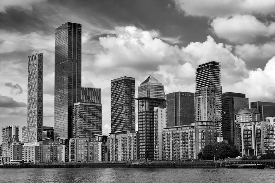 A black and white photograph of a modern city skyline viewed from across a body of water, featuring a range of high-rise buildings with glass facades and varied architectural styles. In the foreground, there are mid-rise residential or commercial structures with balconies and uniform windows, located along the waterfront. The taller skyscrapers in the background include a mix of rectangular, lattice, and geometric designs, with some buildings having visible vertical or horizontal lines and reflective surfaces. The sky above is filled with scattered clouds, casting shadows that highlight the textures and shapes of the buildings. The overall scene captures an urban environment often associated with major European financial districts, where independent or commercial rubbish removal services, such as those provided by Rubbish Clearance Canary Wharf, might operate to support city center waste management, especially in areas with high-density office and residential spaces. The composition emphasizes the contrast between the solid built environment and the dynamic sky, reflecting a typical cityscape scene related to private waste handling and on-site clearance activities.