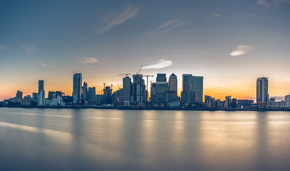 A panoramic view of a modern city skyline featuring numerous high-rise office buildings and skyscrapers with glass facades reflecting sunlight. The cityscape includes construction cranes indicating ongoing development, and the buildings vary in height and architectural style. In the foreground, there is a lush green park with dense, mature trees and well-maintained grassy areas, providing a natural contrast to the urban environment. The sky above is clear with light blue tones and a few wispy clouds, suggesting a bright, sunny day. The scene appears to be taken from a vantage point within the park, showcasing the juxtaposition of natural greenery and city infrastructure, relevant to discussions of urban development, private waste disposal, or site clearance efforts near metropolitan areas, as sometimes handled by independent rubbish removal services or private waste management providers.