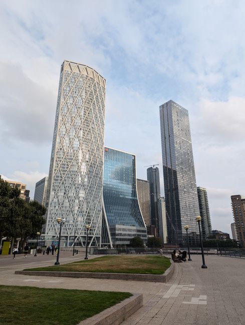 A view of a modern cityscape featuring several tall skyscrapers with glass facades, including a distinctive, lattice-patterned tower on the left, a wider curved glass building in the center, and a taller rectangular tower on the right. The scene is set in a landscaped urban plaza with paved walkways, a small grassy area with a low concrete border in the foreground, and evenly spaced street lamps. The sky above is partly cloudy with patches of blue, creating natural light that reflects off the building surfaces. The area appears to be a public space with minimal human activity, consistent with a contemporary business district focused on office or commercial use, and aligns with the context of non-local authority rubbish management or private waste clearance facilities present in urban development environments.