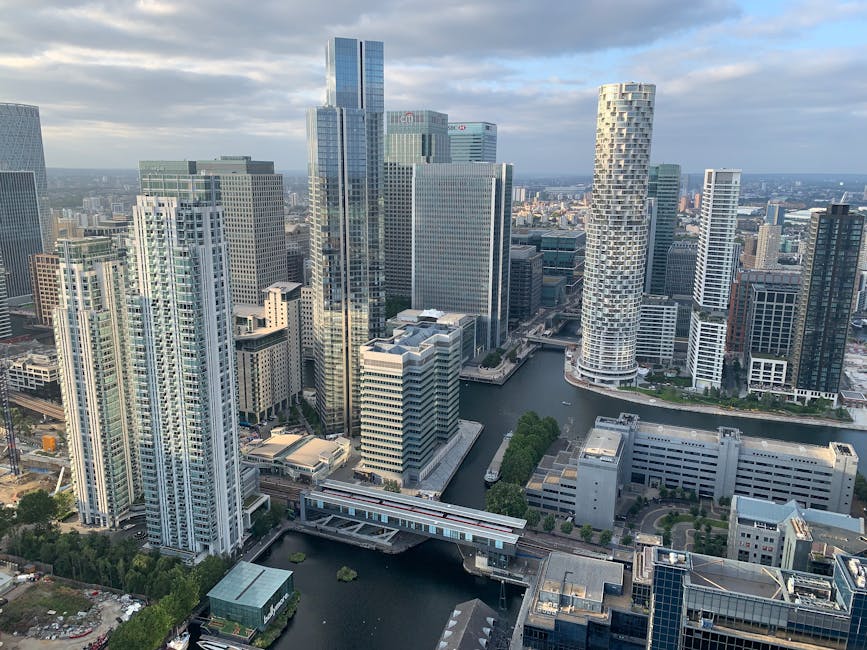 An aerial view of the Canary Wharf financial district featuring numerous modern high-rise office buildings with glass facades, some with reflective surfaces that mirror the sky and surrounding structures. The buildings vary in height, with the tallest ones positioned centrally, and include a distinctive cylindrical skyscraper on the right with a patterned exterior. In the foreground, a canal runs through the area, bordered by concrete pathways and green landscaped sections. The waterway is calm, reflecting parts of the buildings and the cloudy sky above. Smaller commercial and residential buildings occupy the lower areas, with some construction activity visible in the lower left corner. The scene is illuminated by natural daylight, with soft shadows cast by the cloud-covered sky, demonstrating the typical urban environment where private waste or rubbish removal services could be relevant for commercial property maintenance.