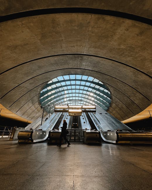 The image depicts the interior of a modern underground station with a high, curved concrete ceiling featuring layered, concentric arches. In the center, there are two escalators with metallic surfaces, leading upwards towards an illuminated glass roof that allows natural light to filter into the space. A person dressed in dark clothing is visible riding the escalators, while another individual wearing similar attire stands nearby, possibly waiting or preparing to ascend. The station's floor is made of smooth, dark stone, and the surrounding walls have a sleek, polished finish, creating a clean and structured atmosphere. The environment appears spacious, with a focus on minimalistic design and functional materials such as concrete, glass, and metal. The scene emphasizes a contemporary transportation hub, aligning with themes of efficient passenger movement and indoor public space, which can relate to the idea of alternative waste handling or facility maintenance within busy urban environments, often managed through independent or private waste removal services by companies like rubbishclearancecanarywharf.co.uk.
