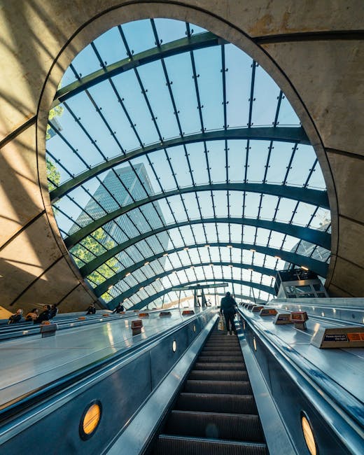 A black and white photograph of an interior view inside a modern train station featuring a large, curved glass and metal ceiling structure allowing natural light to illuminate the space. In the foreground, multiple escalators ascending from the lower level to the upper platform are visible, with several commuters riding or walking up and down. The escalators have black rubber handrails and metal steps, with illuminated signals indicating movement direction. To the right, there is a sign displaying the name 'ADAM' alongside other directional signage. The station's design emphasizes sleek, curved architectural lines with smooth surfaces, creating an open and airy atmosphere. The environment appears clean, well-maintained, and busy with human activity, reflecting a typical scene of a bustling transportation hub in the context of urban infrastructure, indirectly supporting the theme of efficient movement and logistics associated with rubbish removal and waste management within such settings.